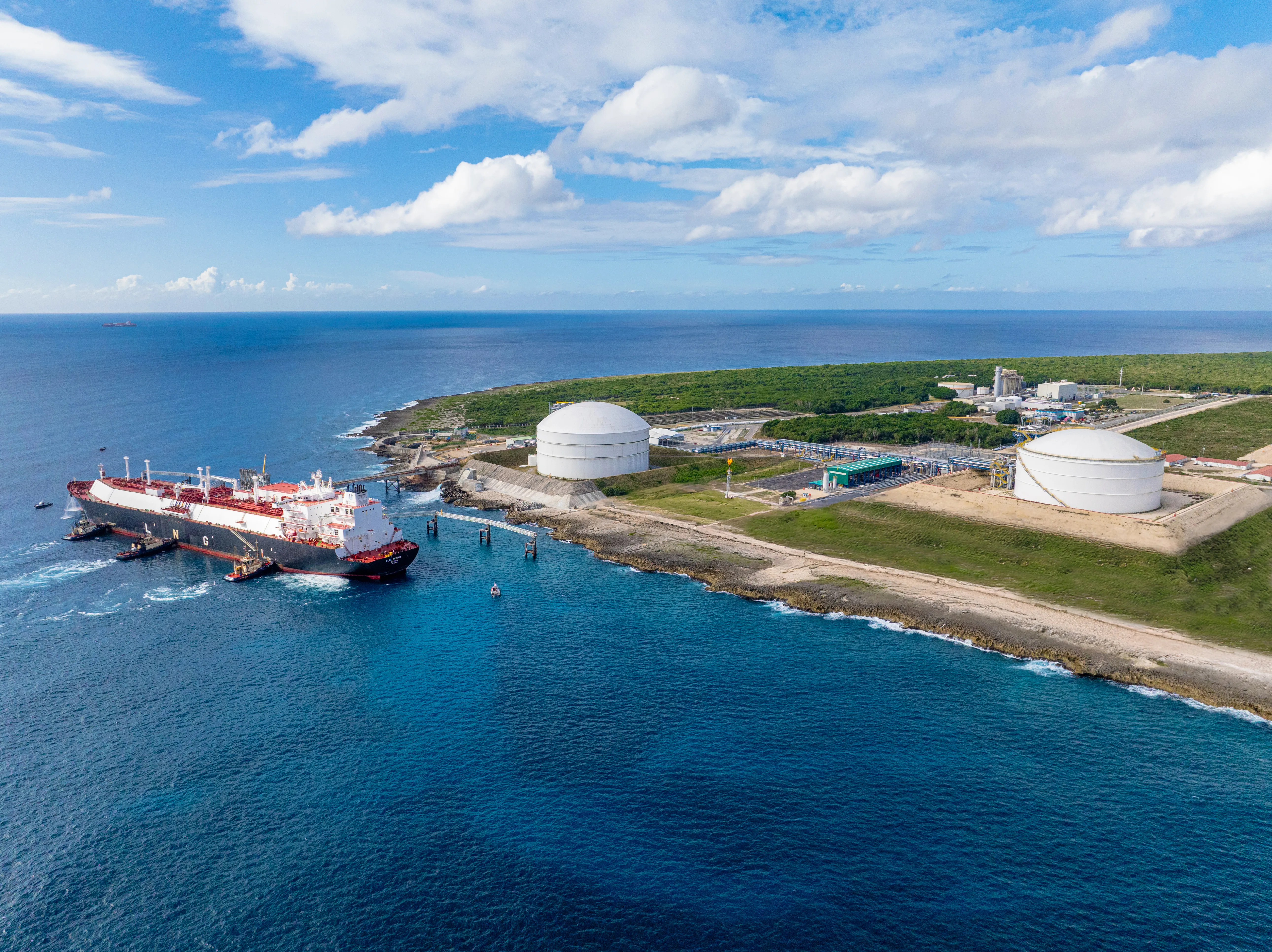 Aerial view of a large tanker ship docked at a coastal LNG terminal with two large white storage tanks. The terminal is situated on a green, grassy landscape next to the ocean under a partly cloudy sky.