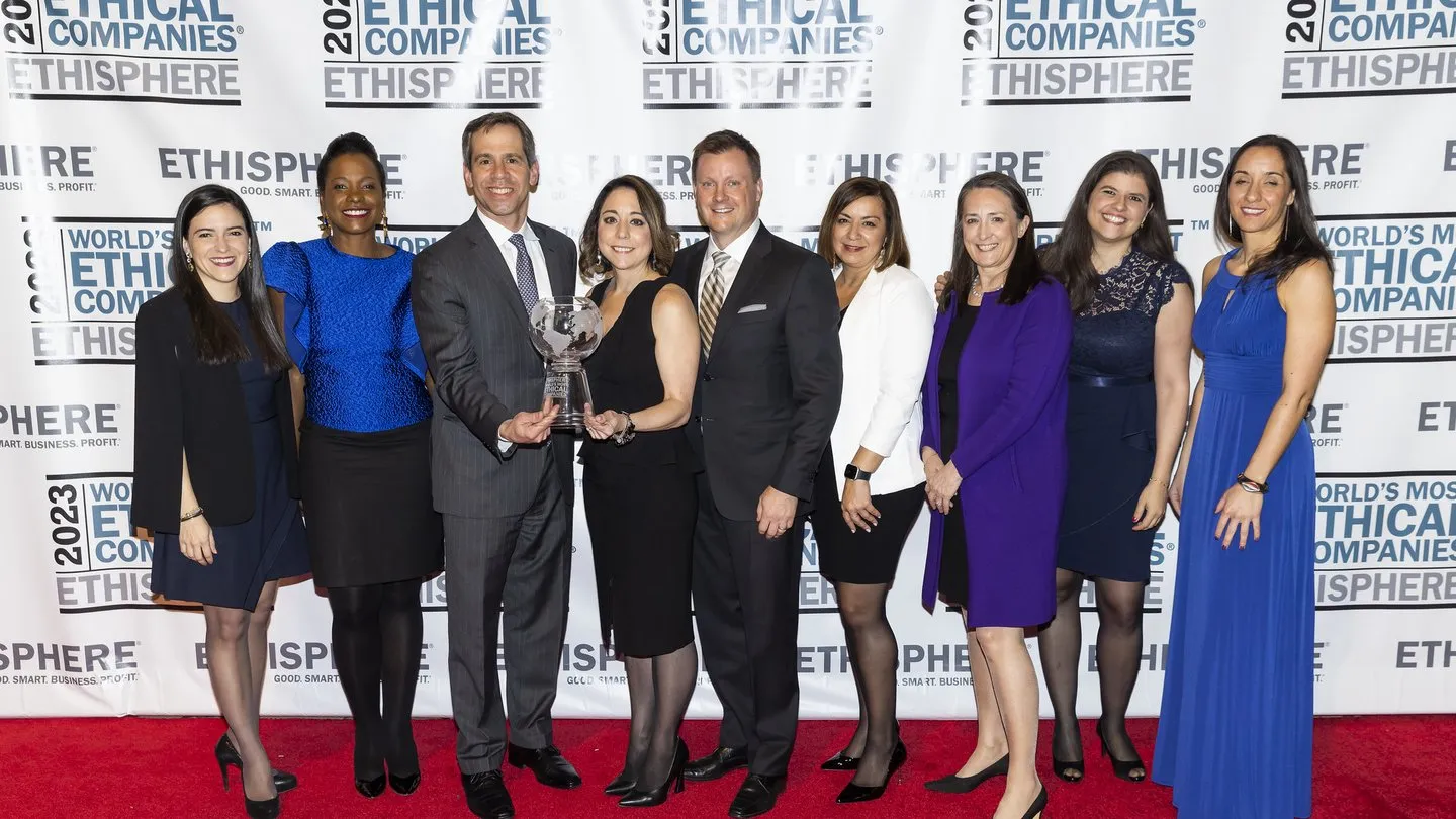 A group of nine people standing on a red carpet, posing with an award. They are dressed in formal attire, with a backdrop displaying 'World's Most Ethical Companies' and 'Ethisphere'.