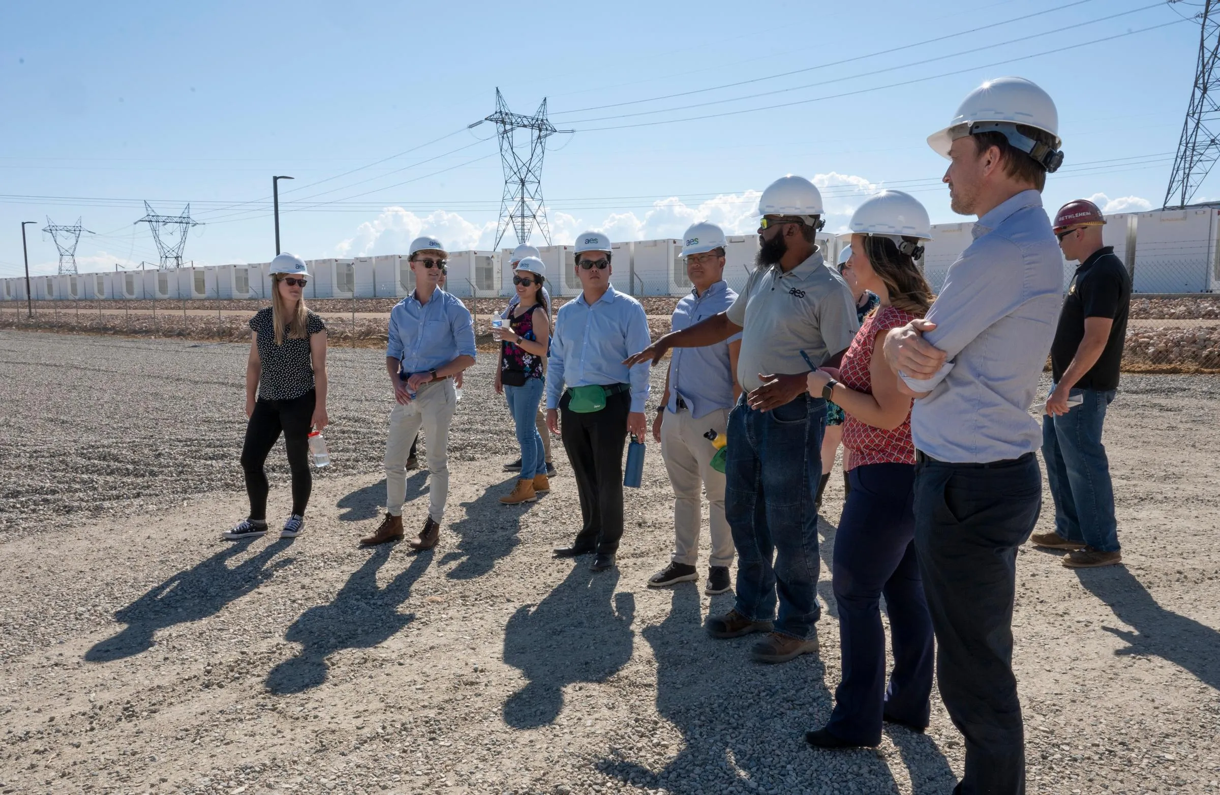 A group of people wearing hard hats and casual clothing stand on a gravel area near electrical infrastructure. They appear to be on a site visit, with power lines and equipment visible in the background under a clear blue sky.