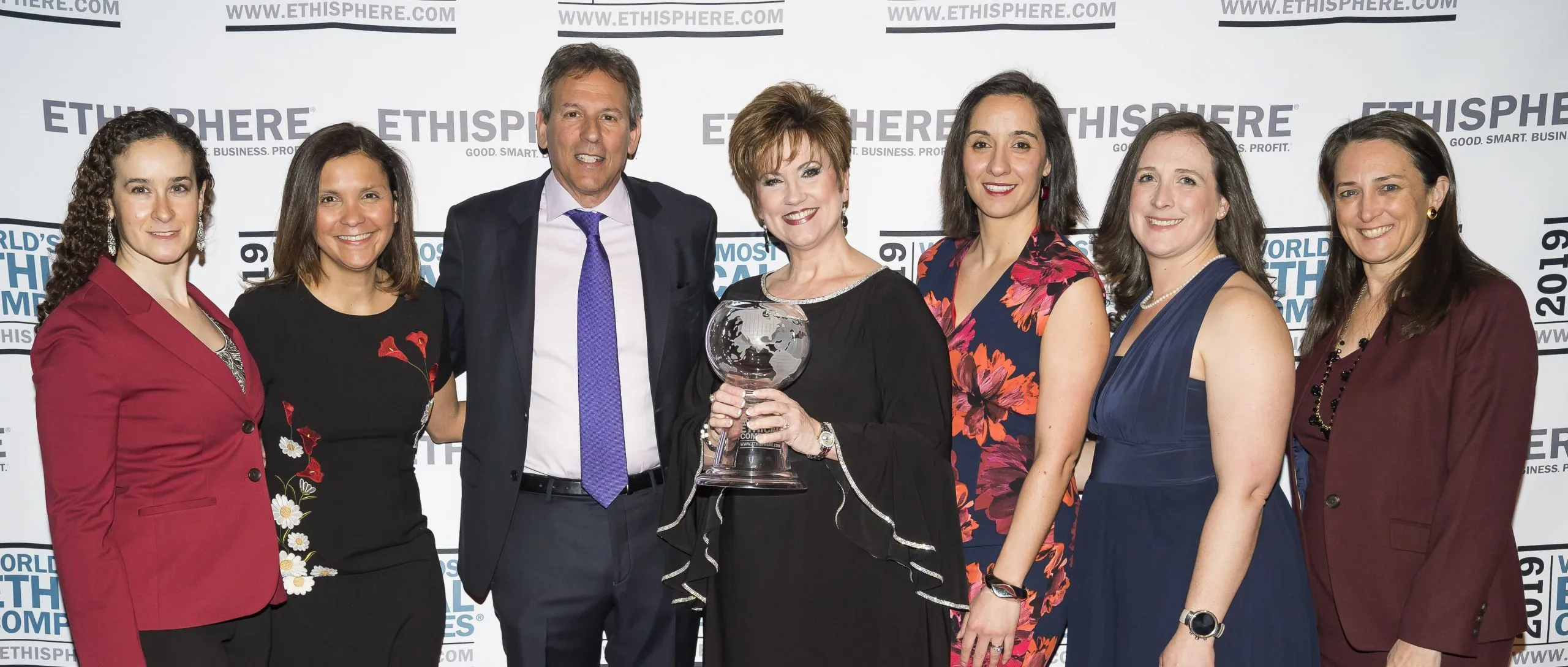 Group of seven people posing for a photo at an event, with a woman holding a glass award. The backdrop displays the Ethisphere logo.