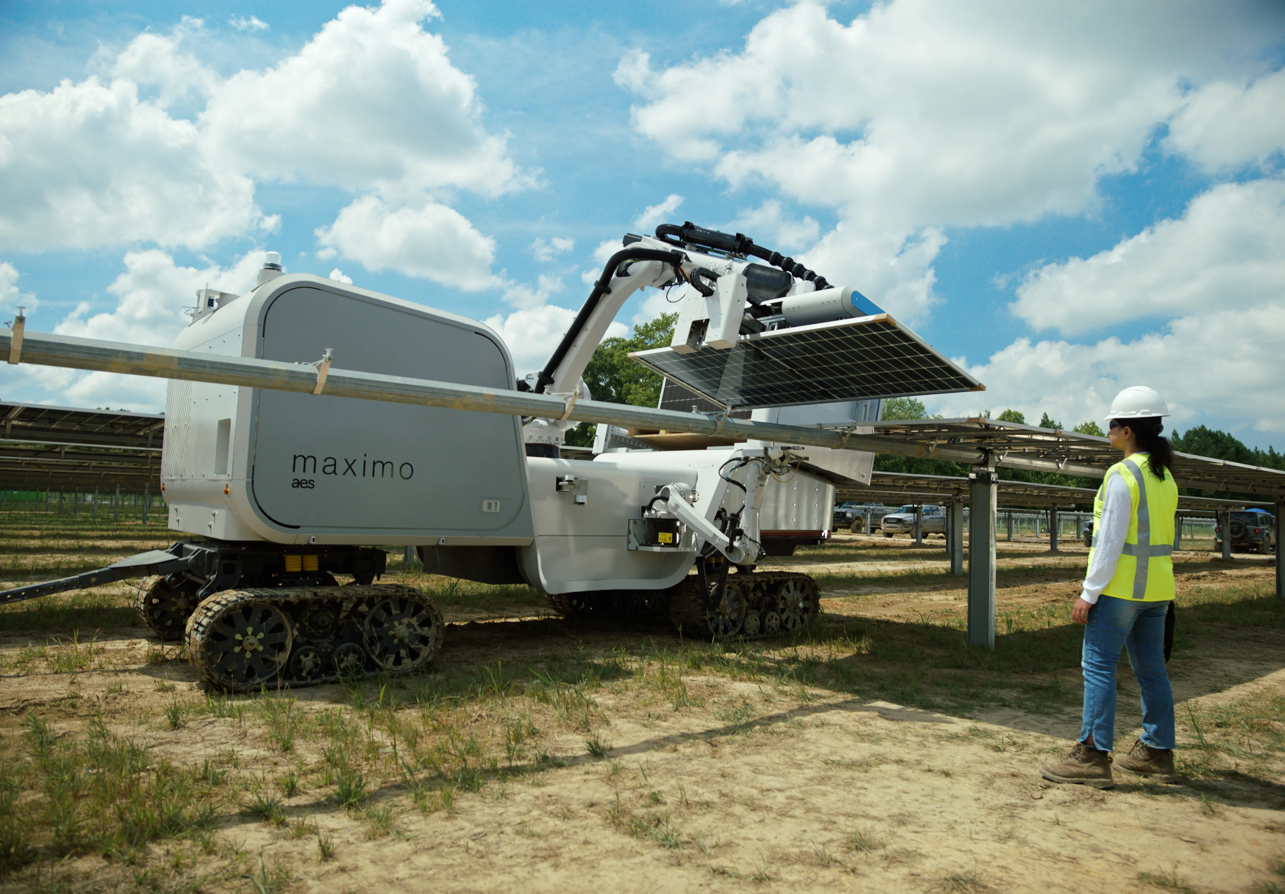 A robotic machine labeled 'maximo aes' is operating in a solar farm, adjusting a solar panel. The machine is on caterpillar tracks, and a person in a safety vest and helmet observes nearby. The background shows rows of solar panels under a partly cloudy sky.
