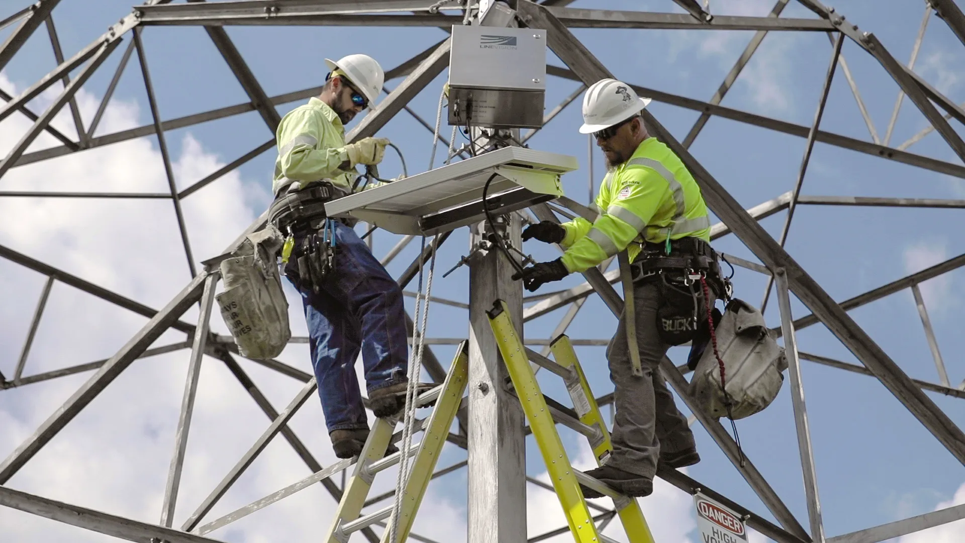 Two workers in safety gear and helmets install equipment on a metal structure. They are on ladders and handling cables, with a clear blue sky in the background.