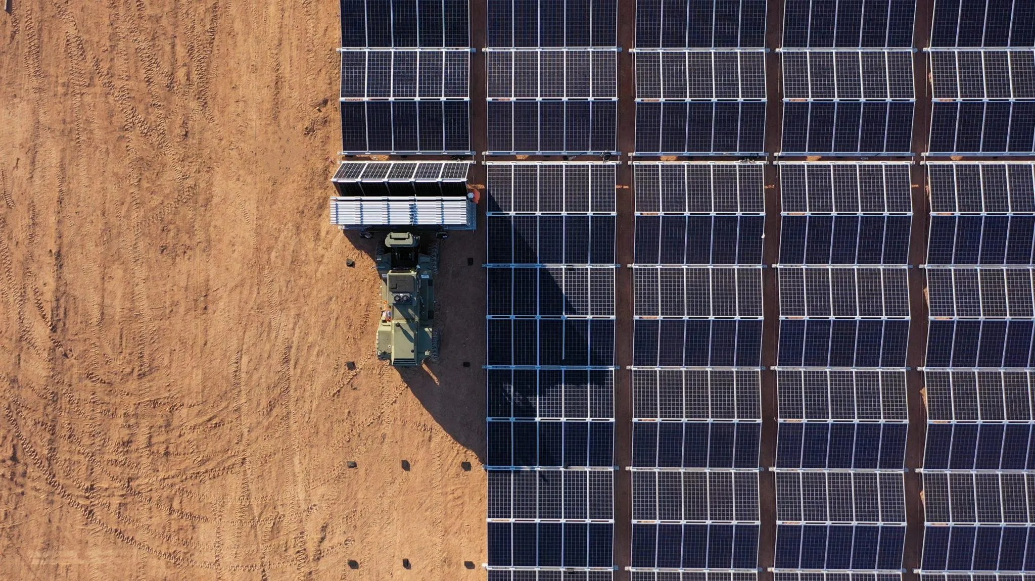 Aerial view of a large solar panel installation on a sandy terrain. A vehicle is parked next to the panels, possibly for maintenance or installation purposes. The panels are aligned in neat rows, casting shadows on the ground.