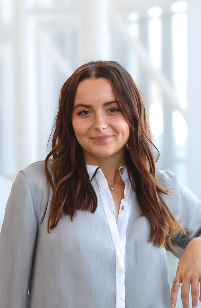 A woman with long brown hair wearing a gray blouse over a white shirt, standing in front of a blurred background with white structures.