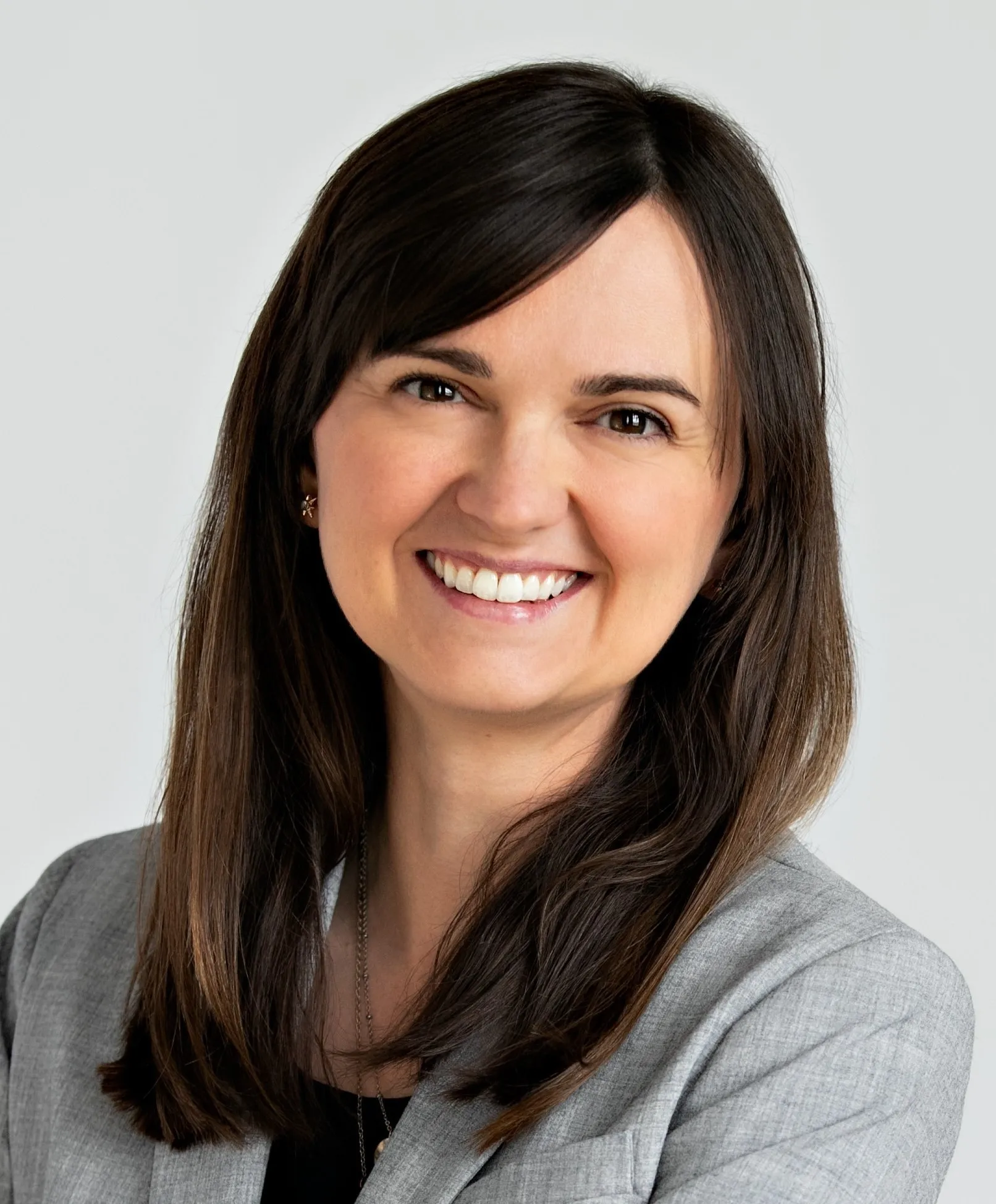 A woman with long brown hair smiling, wearing a gray blazer and a black top against a light background.