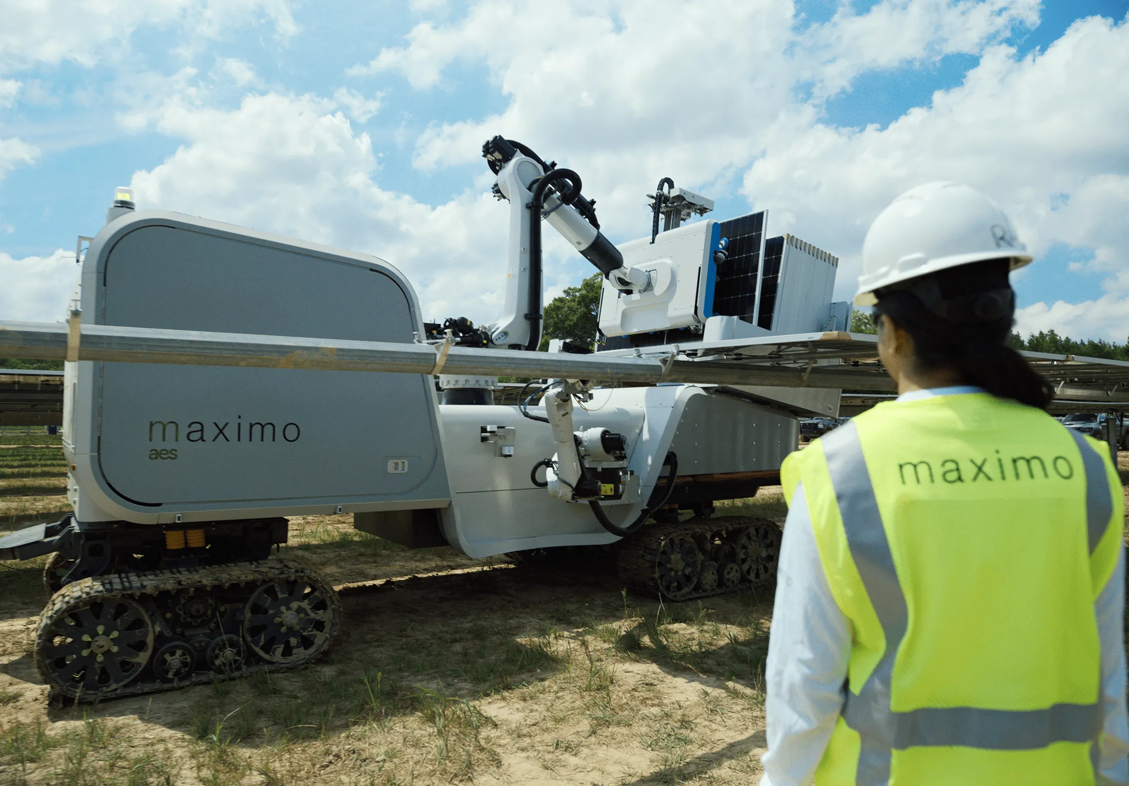 A person wearing a safety vest and hard hat observes a solar panel installation robot labeled 'maximo' on a sunny day.