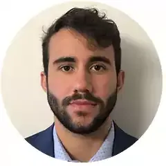 Portrait of a young man with dark hair and beard, wearing a blue suit jacket and light shirt, facing forward against a plain light background.