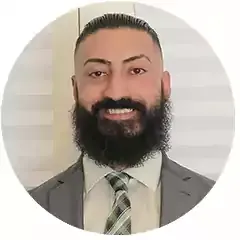 Smiling man with dark hair and full beard wearing a gray suit, light shirt, and striped tie, standing in front of white blinds.