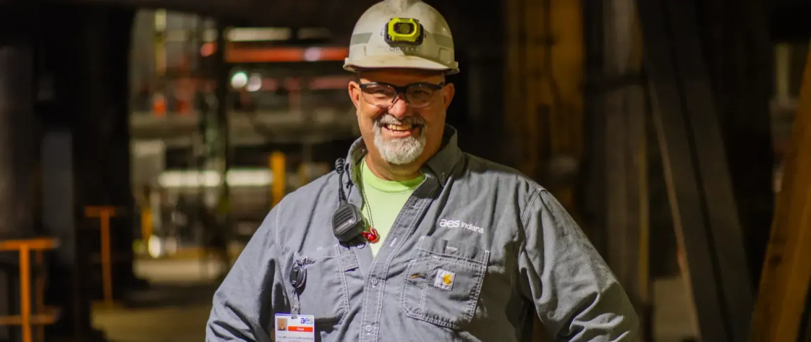 A smiling worker wearing a hard hat and safety glasses stands in an industrial setting. He is dressed in a gray work shirt with a name badge and a communication device clipped to it.