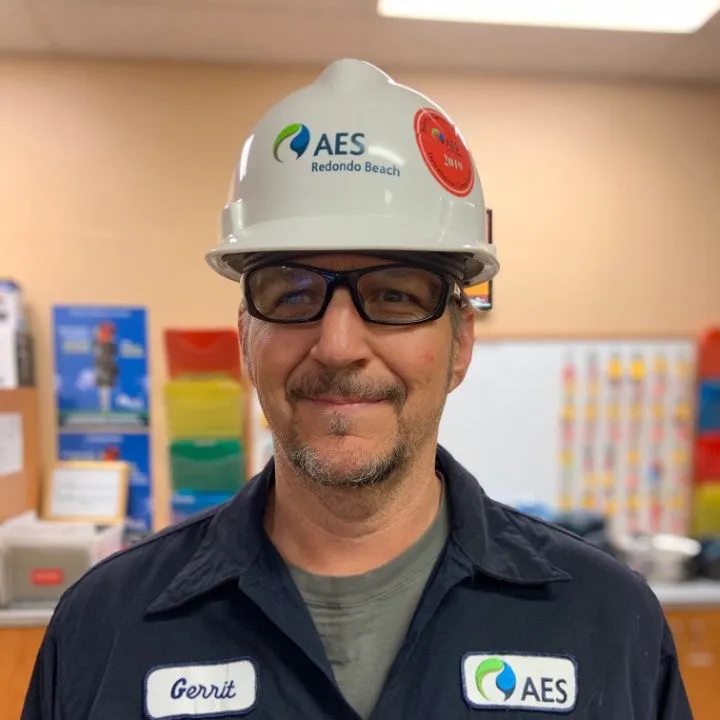 A person wearing a white AES Redondo Beach hard hat and safety glasses, standing indoors with a soft smile. They are dressed in a dark uniform with an AES logo and a name tag. The background is blurred, showing colorful posters and office supplies.