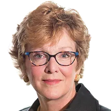 Portrait of a person with short curly hair and glasses, wearing a dark jacket and earrings, looking directly at the camera.