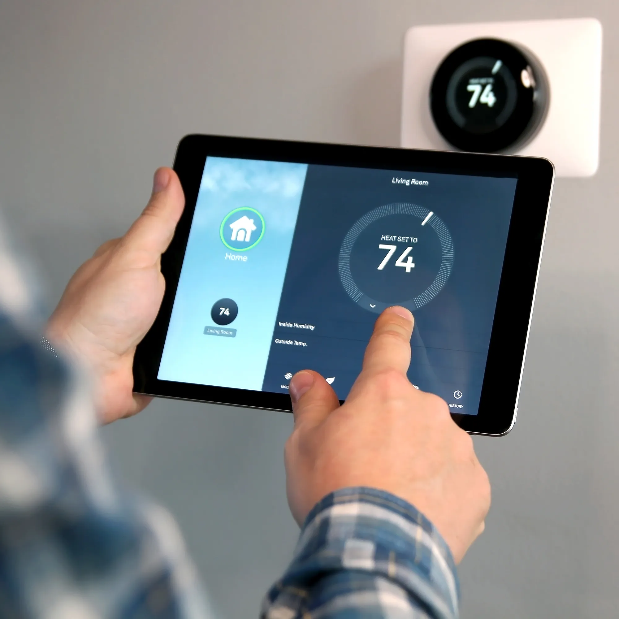 Person adjusting a smart thermostat using a tablet. The screen shows a temperature setting of 74 degrees for the living room. A wall-mounted thermostat displays the same temperature.
