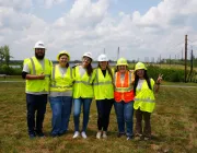 Six people wearing high-visibility vests and hard hats stand together smiling in an open grassy area with solar panels and power lines in the background. The sky is partly cloudy.