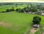 Aerial view of a lush green landscape with open fields, scattered trees, and a few small buildings. A fenced area with cows is visible, along with a dirt road and power lines. The horizon shows rolling hills under a cloudy sky.