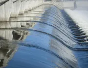 A large dam with water cascading over its edge, creating smooth, wavy patterns. The concrete structure is reflected in the water, and mist rises from the flowing water on the right.