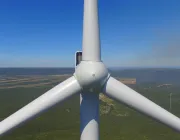 Aerial view of a wind turbine's rotor and blades against a backdrop of green fields and distant hills under a clear blue sky.