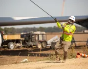 A construction worker wearing a bright yellow safety jacket, hard hat, and gloves pulls on a cable at a work site. In the background, there is a large wind turbine blade, a white truck, and construction equipment on a dirt field.