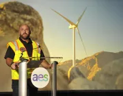 A man in a safety vest speaks at a podium with the AES logo, in front of a backdrop showing a wind turbine and rocky landscape.