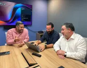 Three men sitting at a conference table, smiling and looking at a laptop. One man is gesturing with his hand. A large screen displaying a meeting schedule is on the wall behind them.