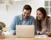 A smiling couple looks at a laptop screen in a bright kitchen, with notebooks and a pen on the table.